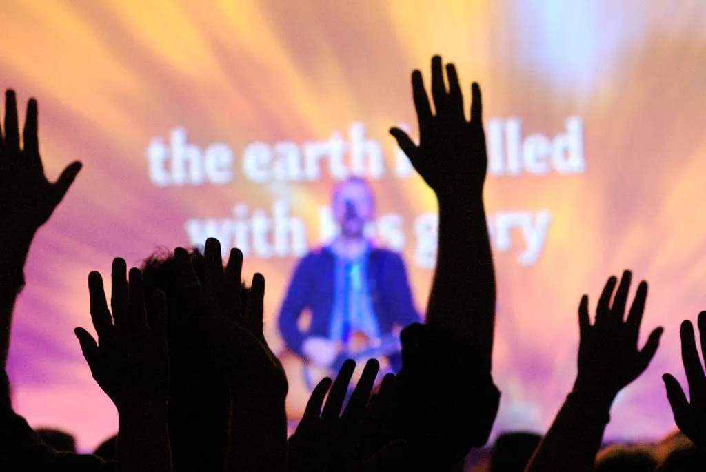 Worshippers raising hands at a concert as lyrics declare "the earth is filled with His glory"