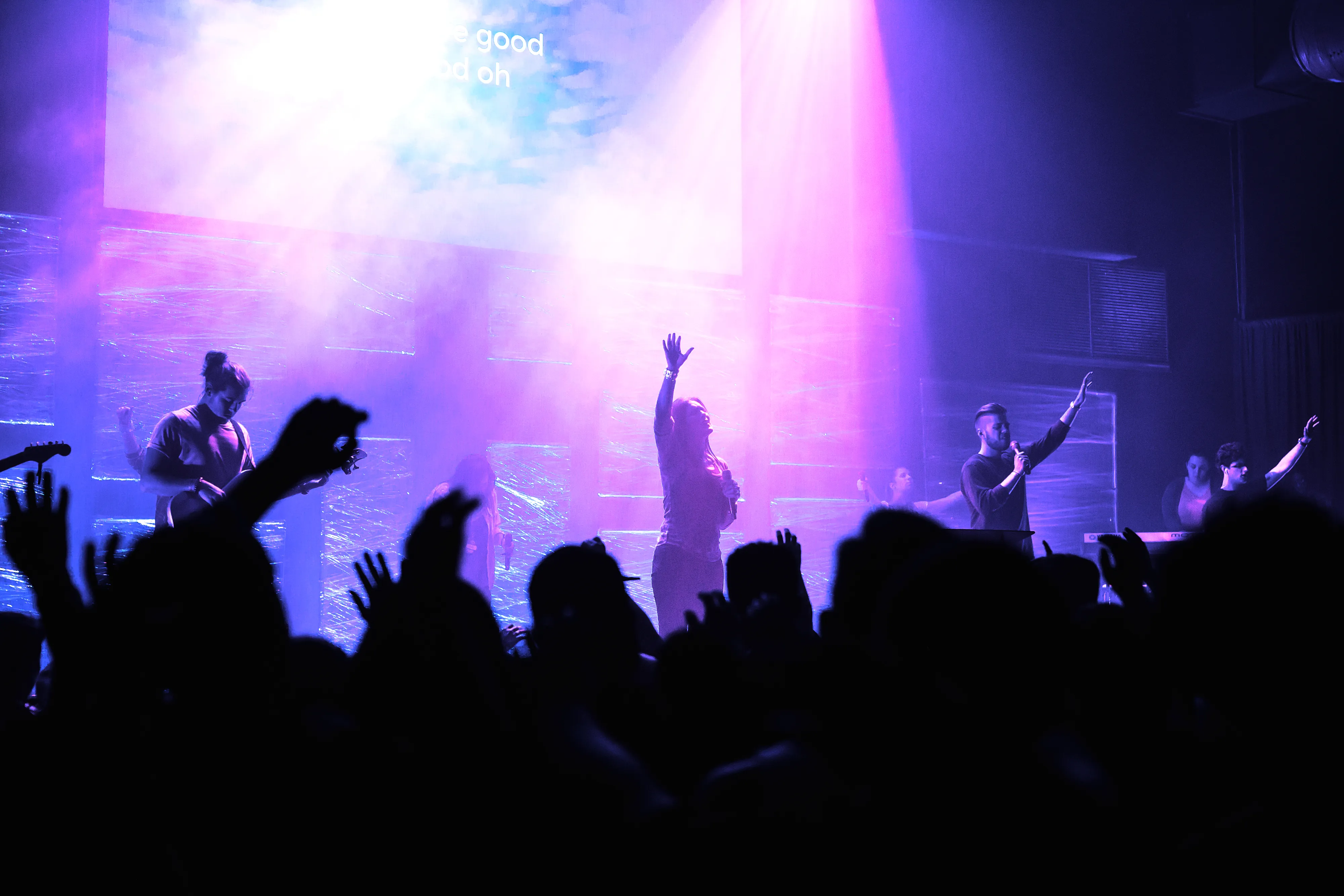 Worship band on a purple-lit stage with the congregation's hands raised